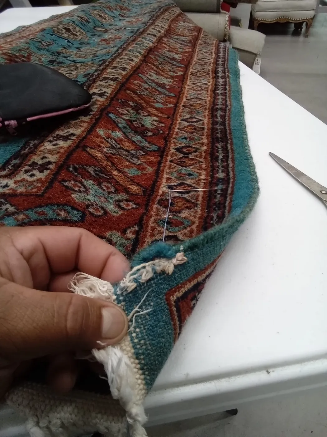 Hands working the fringe of a teal and red rug at the bench